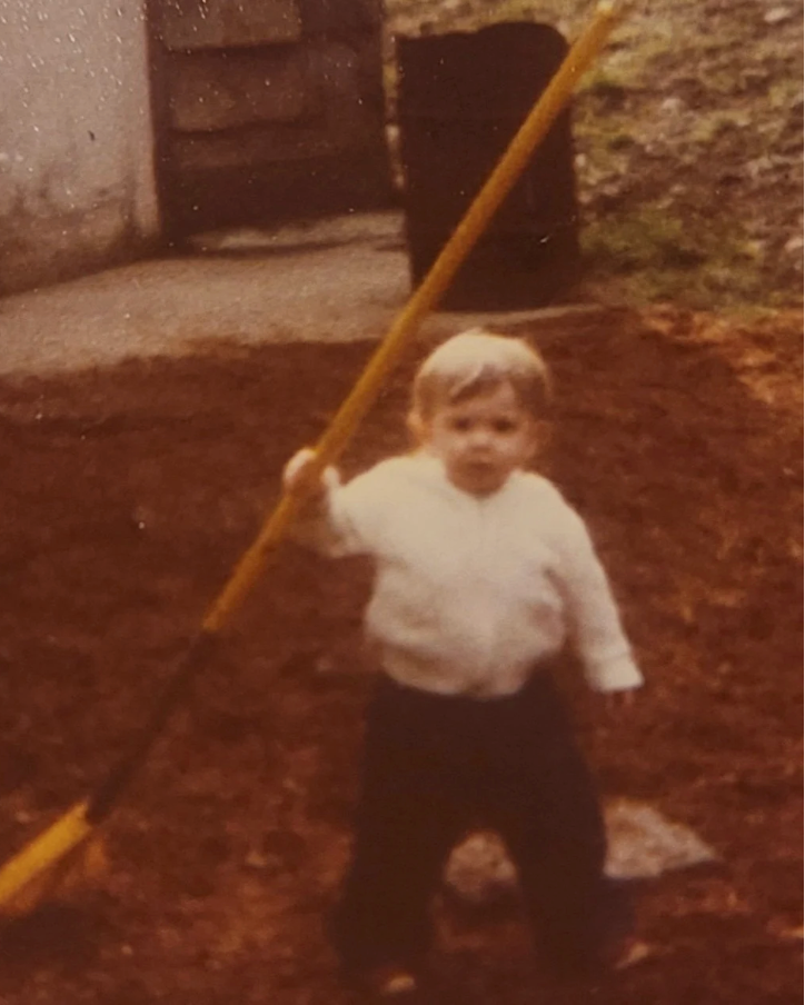 A young Amber Morrison of Bellingham Flower Farm standing outdoors in a yard holding a long yellow-handled gardening tool, with a white shirt and dark pants. The photo has a warm, vintage, sepia-toned quality.