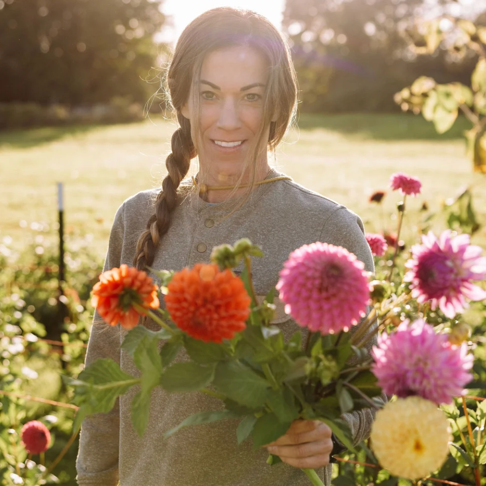 A smiling woman with a braid stands in a sunny flower garden holding a bouquet of colorful dahlia flowers, including pink, orange, purple, and yellow. She is wearing a gray henley shirt.