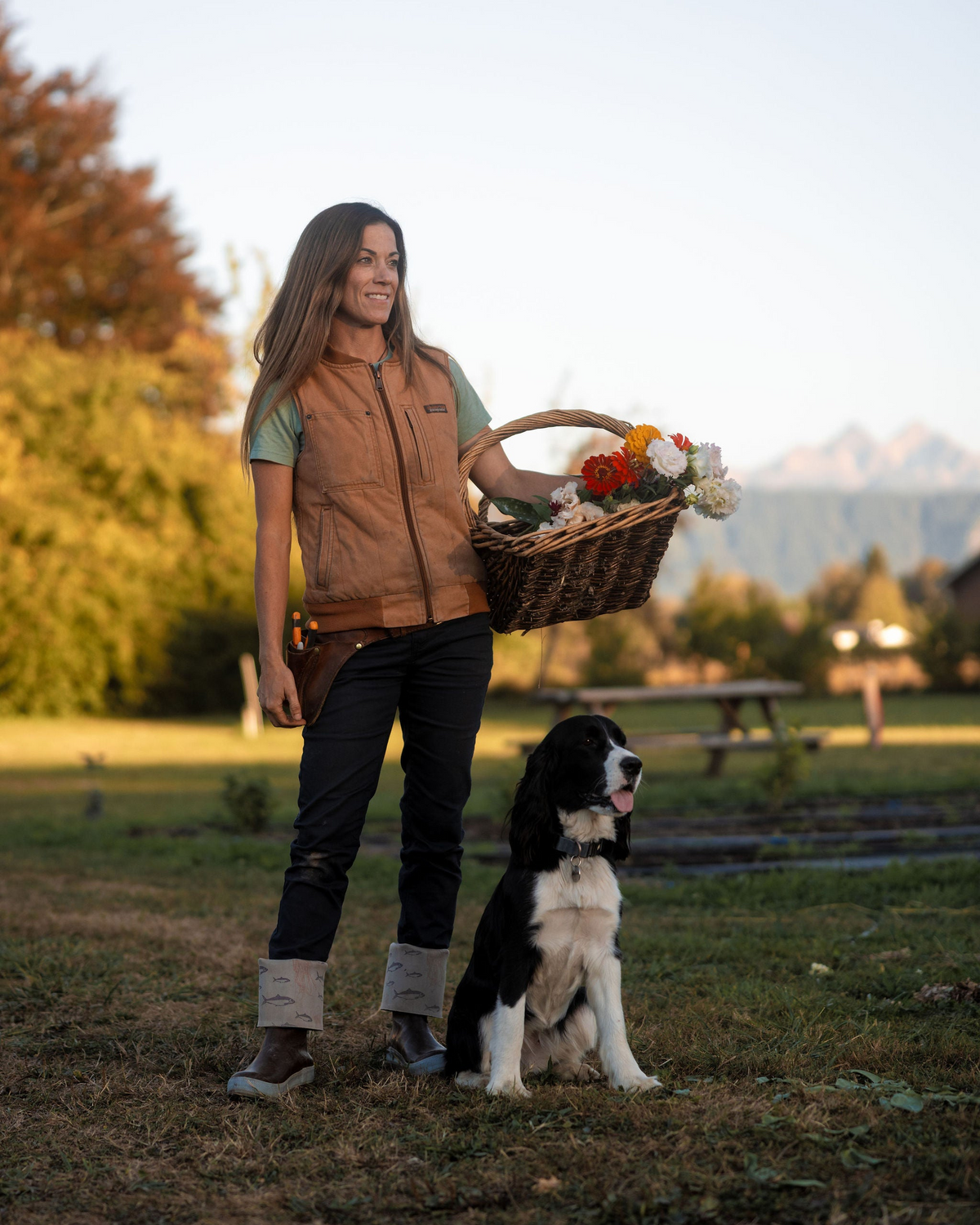 A woman standing outdoors in a grassy field holds a wicker basket filled with flowers next to a black and white dog. She is wearing a brown vest over a green shirt, dark pants, and boots.