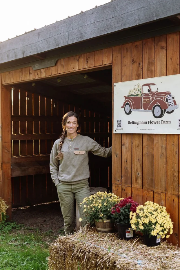 A woman stands in the open doorway of a wooden barn at Bullington Flower Farm, with a sign on the wall showing a vintage pickup truck and the farm name. Hay bales and potted flowers sit in front of the barn on a grassy field.